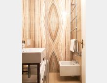 Bagno moderno e compatto con lavandino e bidet sospesi bianchi, parete di fondo rivestita in pietra/marmo venato a motivo simmetrico e scaldasalviette cromato con asciugamano. Pavimento chiaro e porta chiara con maniglia in ottone sul lato destro.