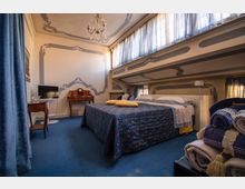 Camera da letto in mansarda con letto matrimoniale, moquette blu e pareti chiare con decorazioni ornamentali grigie. Grandi finestre alte con tende azzurre illuminano l’ambiente; sono presenti lampadario, scrivania in legno e TV su tavolino.