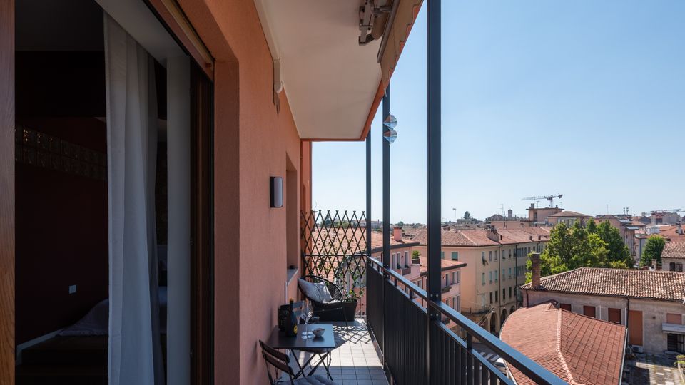 Balcone lungo e stretto di un appartamento con pavimento piastrellato, ringhiera metallica nera e tenda da sole superiore. Sono presenti un piccolo tavolo e sedie pieghevoli e la vista si apre sui tetti in tegole rosse e sugli edifici della città sotto un cielo sereno.