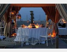 Tavolo rotondo apparecchiato con tovaglia bianca, sedie in ferro battuto e candele, all’interno di un gazebo con tende drappeggiate su una pedana in legno. Sullo sfondo si vede la spiaggia al crepuscolo con lanterne sospese e una palma.