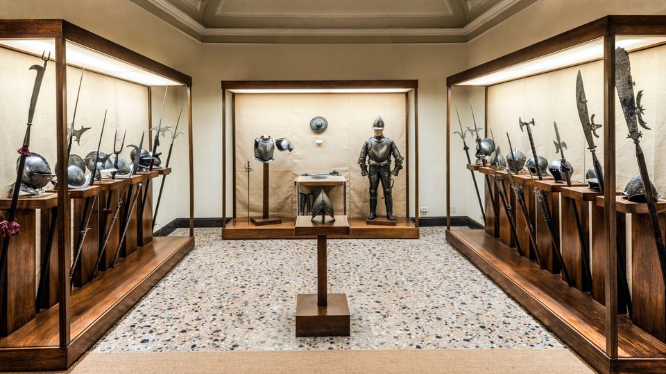 Sala museale con pavimento in seminato e soffitto a volte, con grandi vetrine in legno illuminate che espongono armi antiche e elmi lungo i lati. Al centro c’è una teca con armatura completa e oggetti esposti su piedistalli.