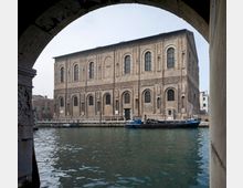 Veduta di un grande edificio storico in mattoni affacciato su un canale, con due piani di alte finestre ad arco e una banchina con pali d’ormeggio. La scena è incorniciata da un arco in pietra in primo piano e sull’acqua è ormeggiata un’imbarcazione.