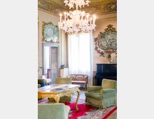 Salone elegante in stile barocco con grande lampadario in vetro, soffitto decorato e pareti ornate da cornici dorate con affreschi. Nella stanza ci sono poltrone verdi, un tavolino intagliato, un pianoforte nero e un’ampia finestra con tende bianche su tappeto rosso.