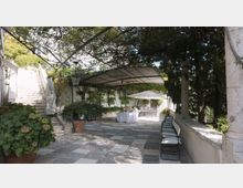 Terrazza in pietra con pavimentazione a lastre, coperta da una pergola con rampicanti e circondata da alberi. Sul fondo ci sono un tavolo apparecchiato sotto un grande ombrellone bianco e sedie allineate lungo un muretto; a sinistra una scalinata in muratura sale verso l’edificio.