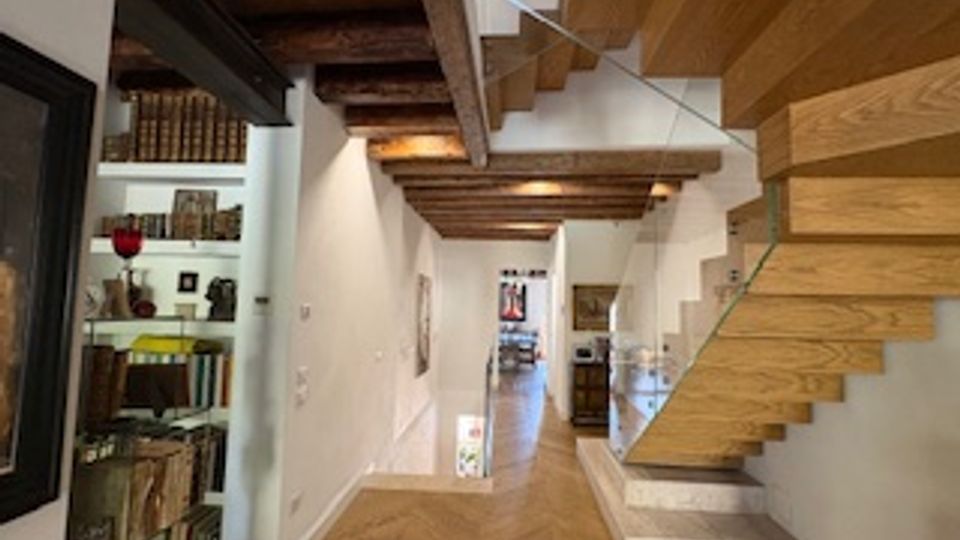 Interno di un’abitazione con pareti bianche, soffitto con travi in legno a vista e pavimento in parquet a spina di pesce. A destra una scala moderna sospesa in legno e, a sinistra, una libreria a tutta altezza con libri e oggetti decorativi.