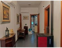 Corridoio interno di un’abitazione con pareti bianche, pavimento scuro e porte in legno che conducono ad altre stanze. Sulla sinistra ci sono quadri e mobili in legno, mentre sulla destra è visibile una stufa a pellet nera e rossa vicino all’ingresso di una stanza.