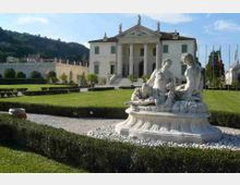 Giardino formale con siepi basse e vialetti di ghiaia, con una grande fontana scultorea in marmo in primo piano. Sullo sfondo si vede una villa neoclassica con portico a colonne e frontone, circondata da prati e colline boscose.