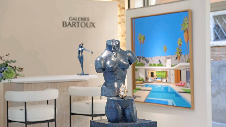 Interno di una galleria d’arte con pareti bianche e insegna “Galeries Bartoux”, bancone con sgabelli e una scultura in primo piano su piedistallo. Sullo sfondo è appeso un grande quadro raffigurante una villa moderna con piscina e palme.