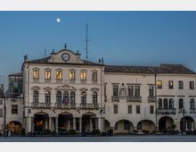 Facciata di un palazzo storico con portici ad archi al piano terra, balconate e finestre simmetriche, sormontata da un frontone con orologio. L’edificio si affaccia su una piazza ampia, con tavolini all’aperto e cielo serale con la luna visibile.