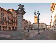 Ampia piazza lastricata con un alto piedistallo in pietra sormontato da una statua alata, circondata da edifici storici con portici e facciate color pastello. Sullo sfondo si vede un campanile in mattoni e, in primo piano, un lampione decorativo in ferro.