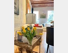 Interno di un soggiorno luminoso con pavimento in parquet a spina di pesce e travi in legno a vista. In primo piano un tavolino metallico con lampada e vaso di tulipani gialli, sullo sfondo un divano chiaro vicino a grandi finestre.