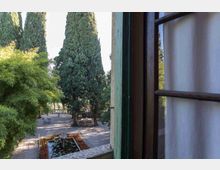 Veduta da una finestra aperta su un giardino con alti cipressi, vialetti in ghiaia e una piccola fontana rettangolare con ninfee. Sullo sfondo si vedono panchine e una balaustra in pietra.