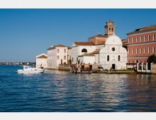 Complesso di edifici storici affacciati su una laguna, con una chiesa bianca dalla facciata barocca e campanile accanto a palazzi dai tetti in coppi. In primo piano si vede l’acqua con un’imbarcazione e un piccolo pontile lungo la riva.