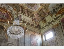 Interno di una chiesa barocca con soffitto affrescato raffigurante colonne e scene allegoriche, decorazioni dorate e un grande lampadario di cristallo sospeso al centro. Un'apertura rettangolare lascia entrare la luce naturale sulla parete laterale.