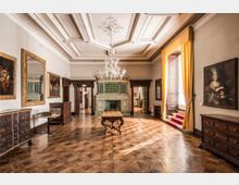 Salone storico con pavimento in parquet a motivi geometrici, soffitto a cassettoni con lampadario in vetro e grande camino rivestito di piastrelle verdi decorate. Alle pareti sono appesi quadri con cornici dorate e sono presenti mobili in legno scuro; a destra una grande finestra con tende gialle e gradini rivestiti di moquette rossa.