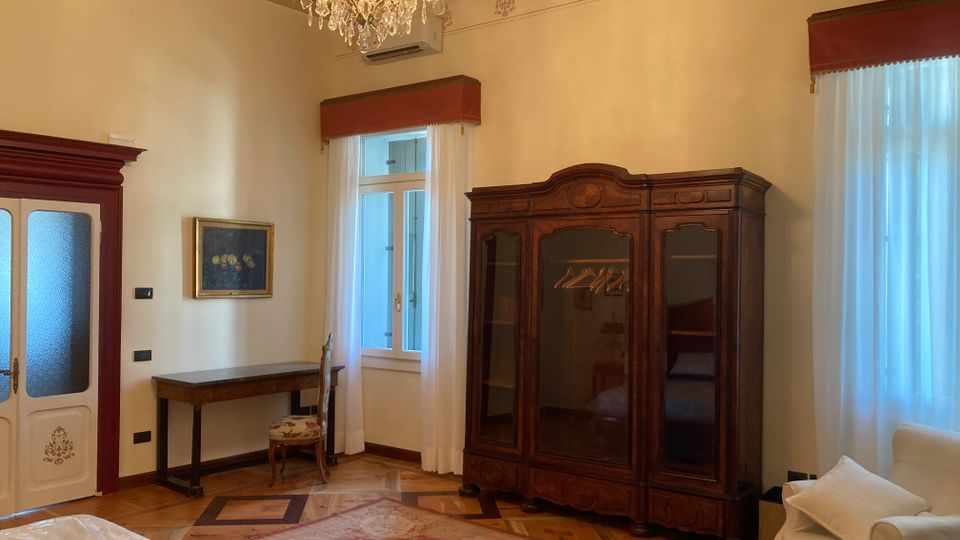 Interno di una camera arredato in stile classico, con armadio in legno scuro, scrittoio con sedia e lampadario di cristallo sotto un soffitto con travi a vista. Due grandi finestre con tende bianche e mantovane rosse illuminano un pavimento in parquet con tappeto e una poltrona chiara.