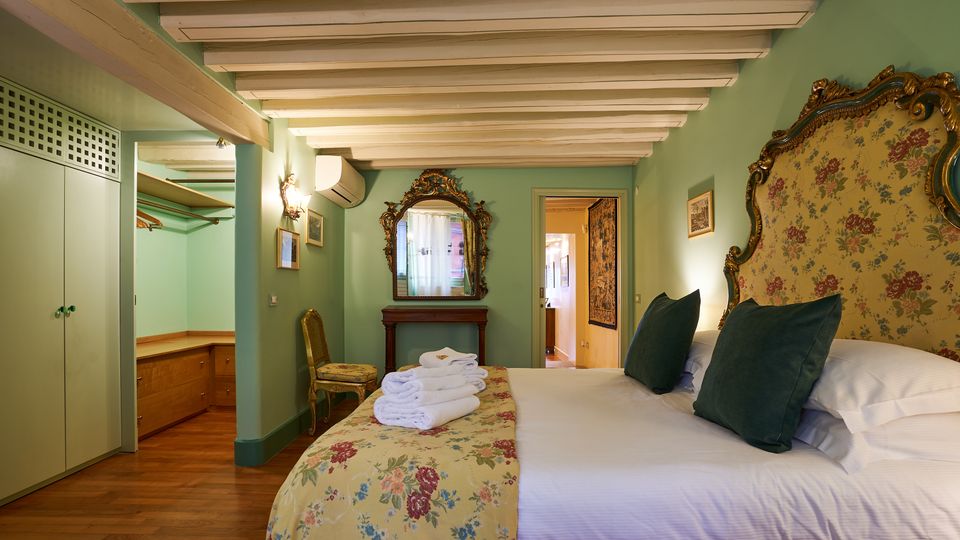 Camera da letto in stile classico con pareti verde chiaro e soffitto con travi a vista bianche, dominata da un grande letto con testiera decorata e copriletto floreale. Nell’ambiente si vedono armadio e cabina guardaroba aperta, specchiera ornamentale su consolle e pavimento in legno.