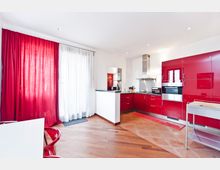 Cucina moderna e luminosa con mobili laccati rossi, elettrodomestici in acciaio inox e piano di lavoro ad angolo. Ampia finestra con tende rosse e bianche, pavimento in legno e piastrelle, illuminazione a faretti incassati.