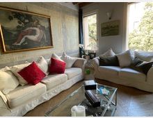 Soggiorno luminoso con due grandi divani chiari e cuscini decorativi, tavolino in vetro e cornice a parete. Finestre ampie con vista sul verde, pavimento in legno e soffitto con travi a vista.