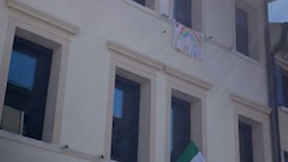 Facciata di un edificio a due piani intonacato di bianco, con grandi finestre rettangolari e un piccolo balcone con bandiera italiana e striscione. In basso si vede un cartello di parcheggio su strada, con cielo sereno sullo sfondo.