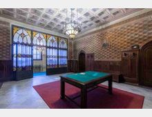 Sala storica con soffitto a cassettoni e lampadario centrale, pareti rivestite in legno e carta da parati a motivo geometrico. In primo piano un tavolo con piano verde su tappeto rosso, sul fondo grandi porte vetrate decorate ad arco che conducono a un corridoio.