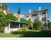 Villa di campagna a più piani con facciata chiara, persiane scure e due camini sul tetto, affacciata su un giardino con prato e alberi. In primo piano una veranda vetrata con colonne e tenda verde, circondata da rampicanti e un grande ombrellone.