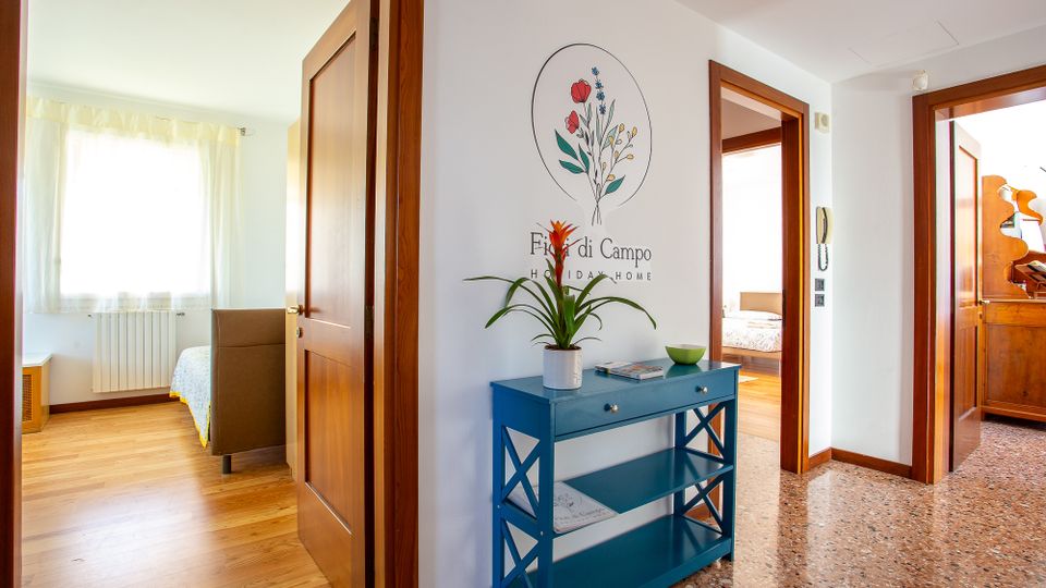 Ingresso di una casa vacanze con pavimento in terrazzo e pareti bianche, con una consolle blu e una pianta sotto un disegno floreale a parete. Porte in legno conducono a camere luminose con parquet e finestra con tende chiare.
