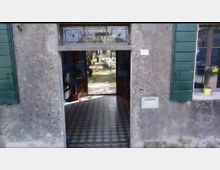 Ingresso ad arco di una casa con facciata in intonaco grigio e persiane verdi, con portone aperto su un corridoio pavimentato a piastrelle. Attraverso il passaggio si intravede un cortile interno con vialetto e vegetazione.