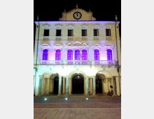 Facciata di un edificio civico storico a tre piani con portico ad archi e colonne al livello stradale, finestre ad arco e balconate con balaustre. In cima è presente un frontone con orologio centrale, illuminato di notte con luci viola e bianche sulla piazza antistante.
