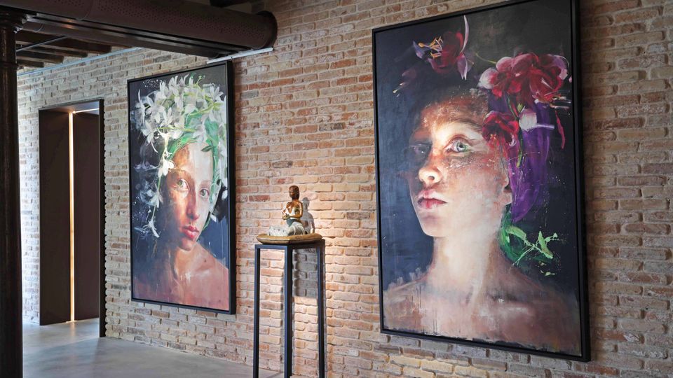 Interno in stile industriale con parete in mattoni a vista, soffitto con travi e condotte, e pavimento in cemento. Due grandi quadri incorniciati appesi alla parete e una piccola scultura su un tavolino alto; a sinistra si vede un corridoio con illuminazione verticale.