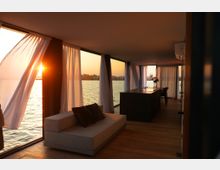 Interno moderno con pavimento in legno e grandi vetrate affacciate sull’acqua, coperte da tende bianche leggere illuminate dal tramonto. In primo piano un divano basso e, sul fondo, un lungo tavolo con sedie vicino alle finestre panoramiche.