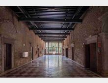 Ampio corridoio interno in un edificio storico con pareti in mattoni a vista e soffitto con travi metalliche, pavimento a scacchi e porte laterali incorniciate in pietra. In fondo, una grande vetrata lascia entrare luce naturale e mostra un’area verde esterna.