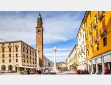 Piazza urbana ampia circondata da edifici storici con portici e facciate colorate, con una torre in mattoni alta con campanile e una colonna monumentale al centro. Sullo sfondo si vedono strade pedonali e un cielo azzurro con nuvole leggere.