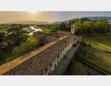 Veduta aerea di un complesso storico in pietra con lunghi tetti in coppi e un campanile, circondato da alberi e campi coltivati. Sullo sfondo si vede un fiume che serpeggia nella valle e una catena di colline e montagne.
