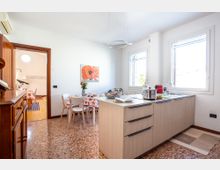 Cucina-soggiorno luminosa con isola moderna in legno chiaro e piano cottura, lavello sotto grandi finestre e pavimento in terrazzo. Sullo sfondo un tavolo da pranzo con sedie bianche, quadro a parete e passaggio verso un altro ambiente con pavimento in legno.