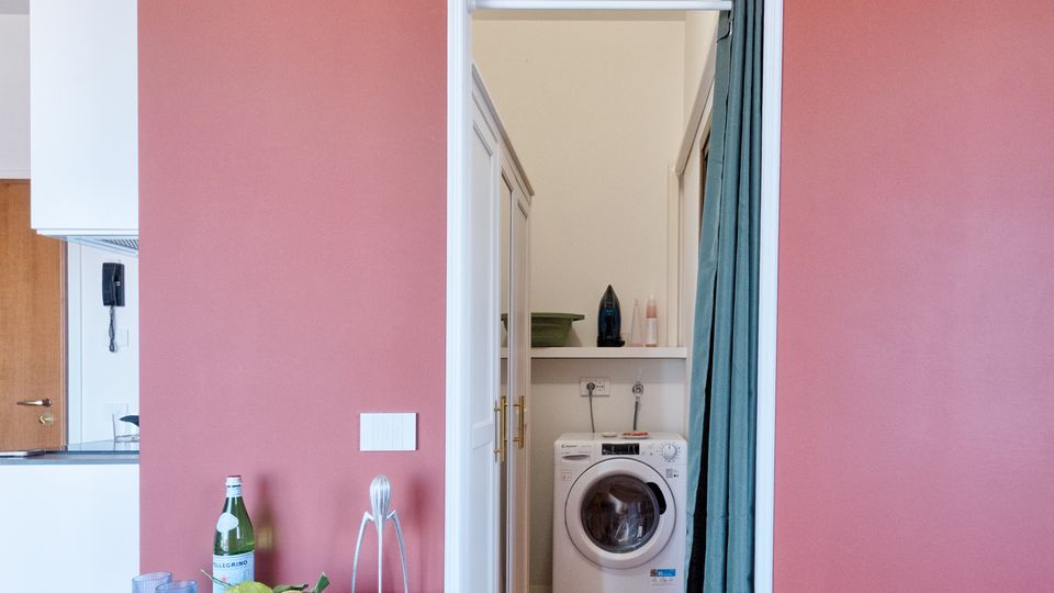 Interno domestico con parete color terracotta e condizionatore a parete sopra una porta che conduce a un piccolo locale lavanderia con lavatrice e mensole. In primo piano pavimento in legno, un mobiletto vintage con oggetti e una scala laterale con inserti in vetrocemento.