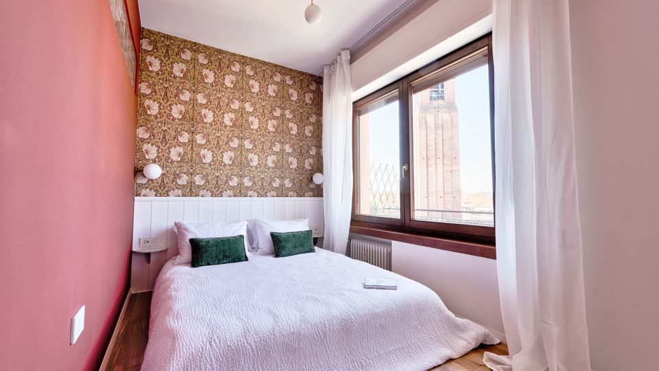 Camera da letto luminosa con letto matrimoniale, parete d’accento rossa e carta da parati decorativa dietro la testata. Ampia finestra con tende bianche e davanzale in legno, con vista su una torre in mattoni all’esterno.