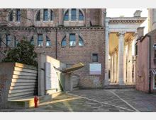 Piccola piazza lastricata tra edifici storici, con una facciata in mattoni e finestre ad arco sullo sfondo. A destra si vede un portico con alte colonne classiche e gradini d’ingresso, mentre a sinistra c’è una struttura moderna in cemento e metallo vicino a un idrante rosso.