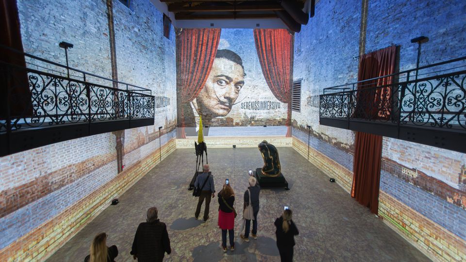 Ampia sala espositiva in un edificio industriale riconvertito, con pareti in mattoni a vista e due balconate laterali in ferro battuto sotto un soffitto con travi in legno. Sul fondo una grande proiezione murale con un volto incorniciato da tende rosse, con sculture esposte al centro dello spazio.