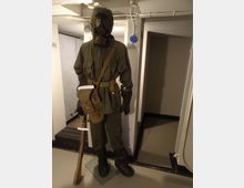 Interno di un bunker o locale sotterraneo con pareti e travi bianche, illuminazione artificiale e un’apertura con scala in discesa. In primo piano è esposto un manichino con equipaggiamento militare e maschera antigas su una pedana, accanto a un fucile.