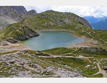 Lago alpino di montagna con acqua turchese, circondato da pendii erbosi e rocce grigie. Sentieri sterrati percorrono le sponde e salgono sulle colline, con cime rocciose e valle sullo sfondo.