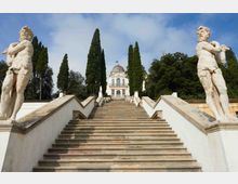 Scalinata monumentale in pietra con parapetti bianchi, fiancheggiata da due statue e da alti cipressi. In cima si vede una villa storica con facciata simmetrica e piccola cupola, circondata da alberi sotto un cielo parzialmente nuvoloso.