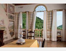 Interno di una sala in stile storico con affreschi alle pareti, soffitto con travi in legno e un tavolo al centro con una grande lampada decorativa. Ampie porte-finestre ad arco con tende bianche si aprono su un balcone, con vista su colline verdi e campagna circostante.