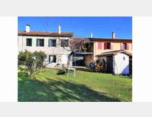 Cortile erboso di una casa rurale con edifici in pietra e intonaco a due piani, tetti in coppi e persiane colorate. Sul prato si vedono piccoli alberi e un’area con tavolo, con un portico e annessi sul lato destro sotto un cielo sereno.