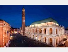 Veduta serale di una grande piazza urbana con un edificio storico a portici su due livelli e tetto verde, affiancato da un alto campanile in mattoni con orologio. Sullo sfondo si vedono altri edifici e l’ampio spazio pavimentato della piazza illuminato da luci artificiali.