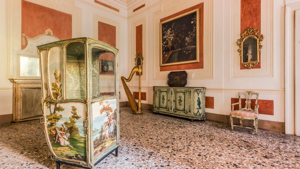 Sala d’interni in stile storico con pareti bianche e riquadri rosso mattone, pavimento in graniglia e arredi antichi. In primo piano c’è una grande alcova paravento dipinta, accanto a un’arpa, una credenza decorata, specchi con cornici dorate e una sedia imbottita.