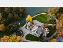 Veduta aerea di una villa storica con tetti in tegole e piccole torrette, circondata da sentieri curvi, prati curati e alberi dai colori autunnali. Accanto si vede un lago o canale che costeggia il parco e una piccola fontana in un’area pavimentata.