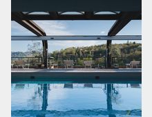 Piscina coperta con ampia struttura in metallo e grandi aperture vetrate sul soffitto, affacciata su una terrazza con ringhiera e lettini. Sullo sfondo si vedono colline alberate sotto un cielo sereno.