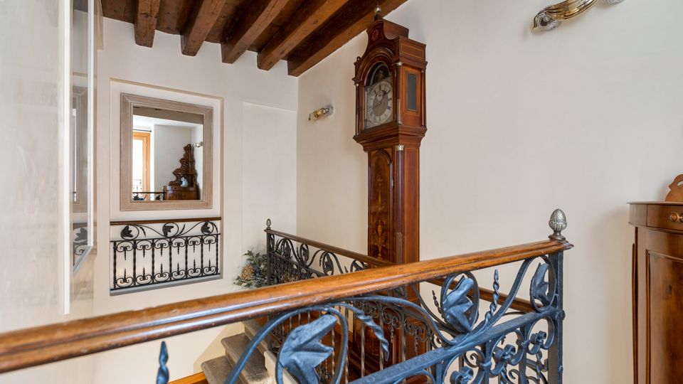 Pianerottolo interno con scala, soffitto a travi di legno a vista e pareti chiare. Ringhiera in ferro battuto decorato, grande orologio a pendolo a parete e passaggio verso un’altra stanza con specchio incorniciato.