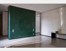 Interno moderno e minimale con pavimento lucido chiaro, grande parete centrale verde scuro arrotondata e pareti in pietra chiara. A destra c’è un lungo piano nero tipo mensola con alcune stoviglie, illuminato da luce naturale che crea strisce sul pavimento.